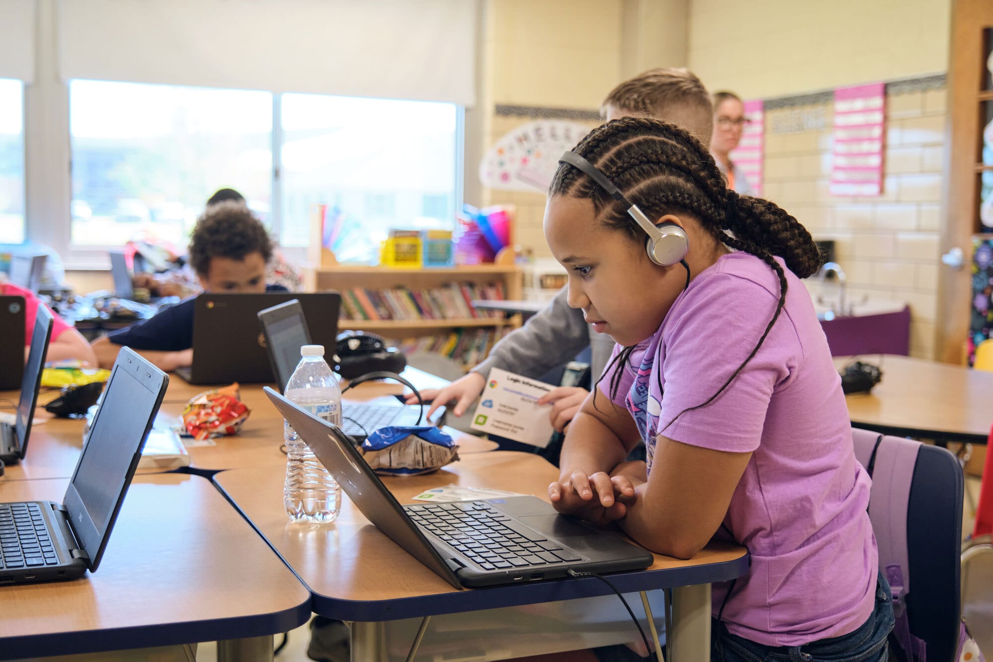 A young girl sits at a classroom desk using a laptop and wearing headphones, with other students working on laptops in the background.