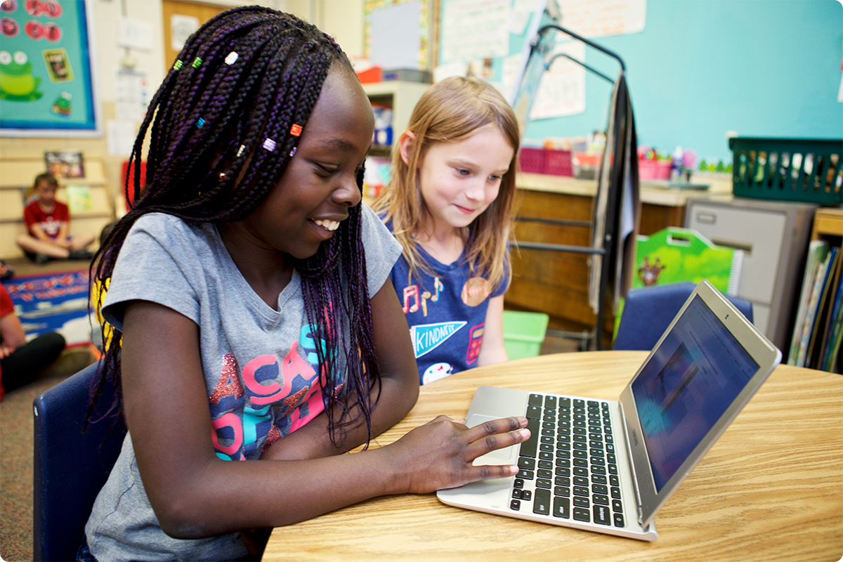 Two young girls, one with braided hair, engaging with a laptop in a colorful classroom setting.