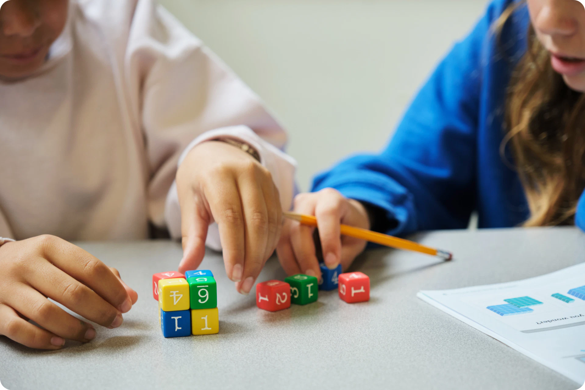 Two children seated at a table engage with colorful number cubes and a pencil beside a worksheet—a delightful way to explore math. This playful setup subtly acts as an informal dyscalculia screener, helping spot characteristics of dyscalculia in young learners.