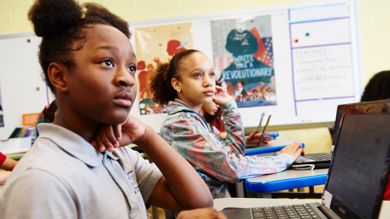 Two schoolgirls sitting in a classroom with laptops, one looking thoughtful and the other looking towards the teacher.