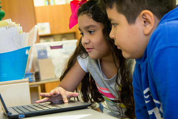 Two children, a girl and a boy, are intently looking at a laptop screen in a classroom setting. the girl is pointing at the screen.