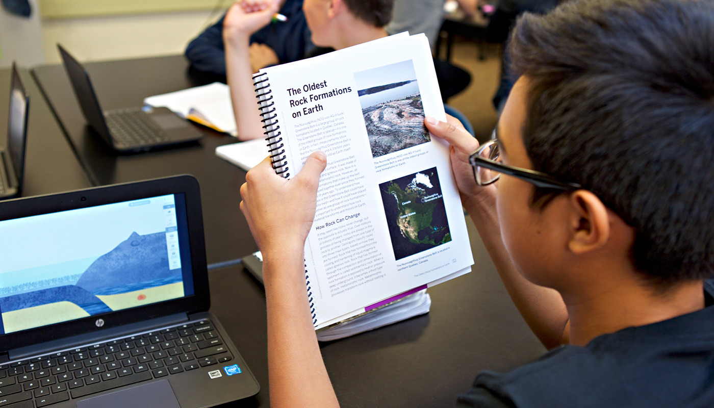 A 鶹holding a textbook about rock formations while using a laptop in a science classroom.