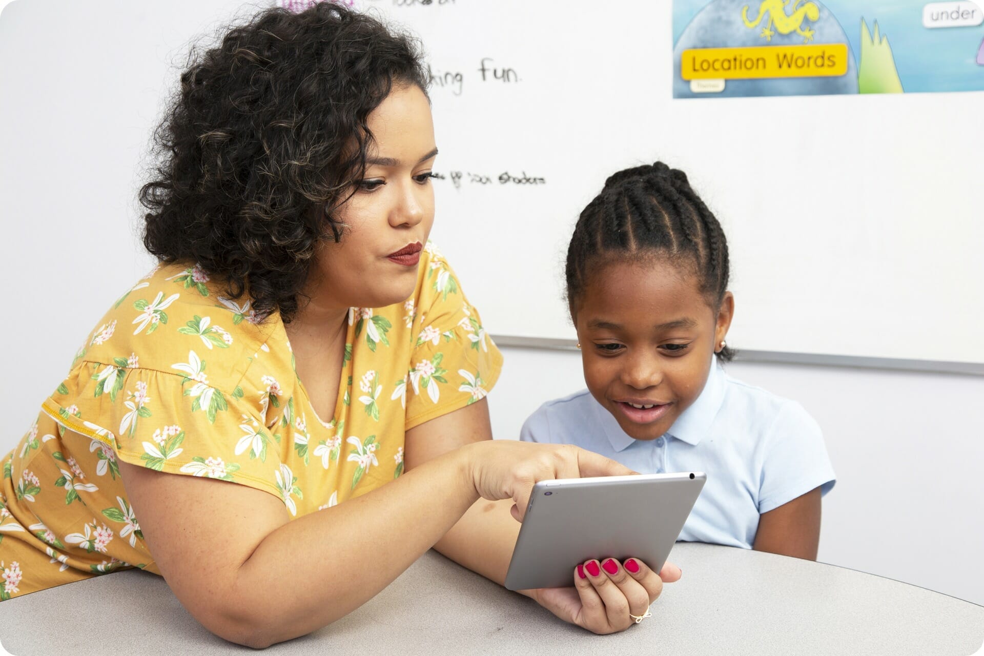 A teacher and young girl focused on a tablet, sitting at a math classroom table, with educational posters in the background.