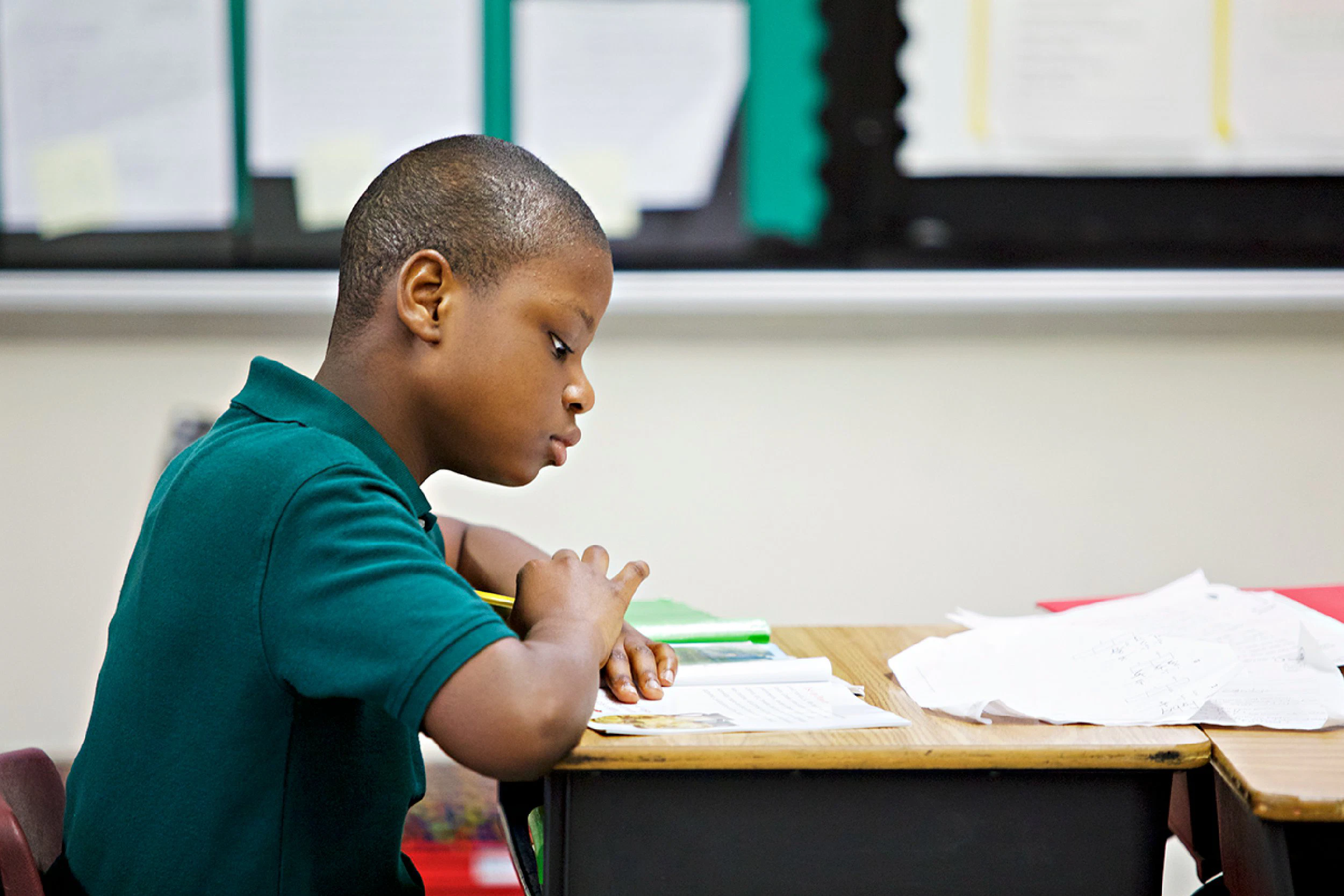 A young boy focusing intently on decoding and phonics as part of his schoolwork at a desk cluttered with papers in a classroom setting.