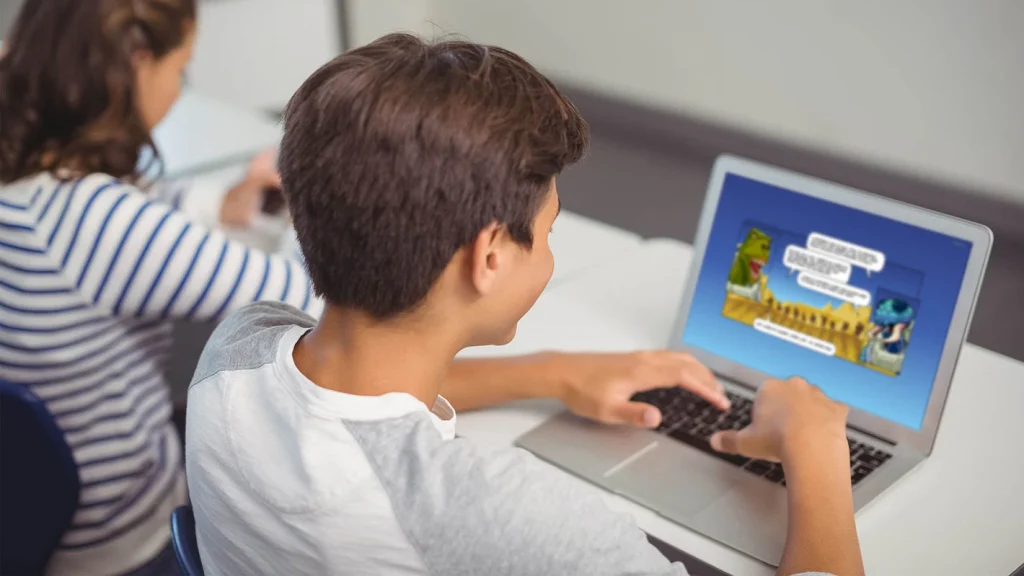 Two children in a classroom, with a boy using a laptop featuring Amplify Reading content on its screen.
