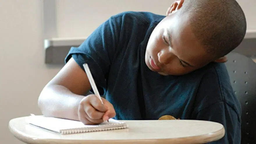 Un niño con la cabeza rapada está sentado en un escritorio, escribiendo en un cuaderno con un bolígrafo blanco.