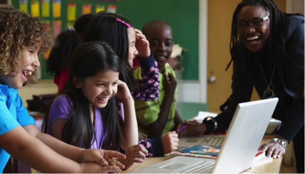 Un grupo de niños y un maestro están reunidos alrededor de una mesa, sonriendo y interactuando con una computadora portátil en un salón de clases.