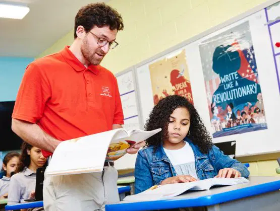 Una maestra con una camiseta polo roja ayuda a una estudiante con su lectura en un salón de clases. Otros estudiantes están sentados en pupitres al fondo. En las paredes hay carteles con mensajes motivadores.