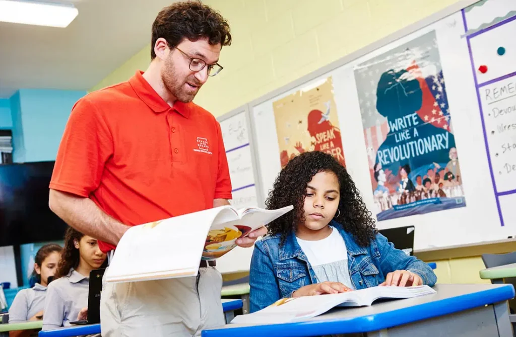 Una profesora con un polo rojo instruye a un alumno de pelo rizado que está leyendo en su escritorio. Otros estudiantes son visibles al fondo, y carteles educativos cuelgan en la pared detrás de ellos.