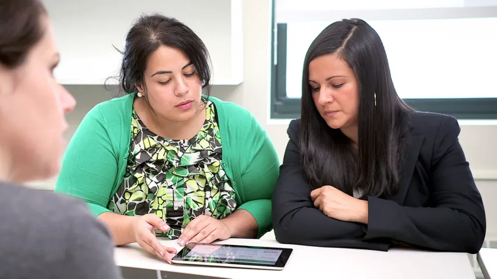 Three women sitting at a table using a tablet to discuss stimulus funding for schools in a professional meeting.