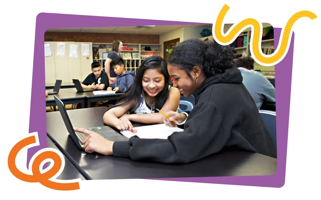 Two students collaborating on a project using a laptop in a classroom filled with other students working in the background.