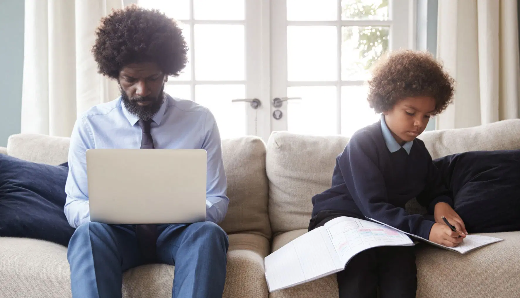 Un adulto y un niño se sientan en un sofá beige con cuadernos abiertos. El adulto usa una computadora portátil, mientras el niño escribe en un libro. La luz del sol entra por las ventanas detrás de ellos.