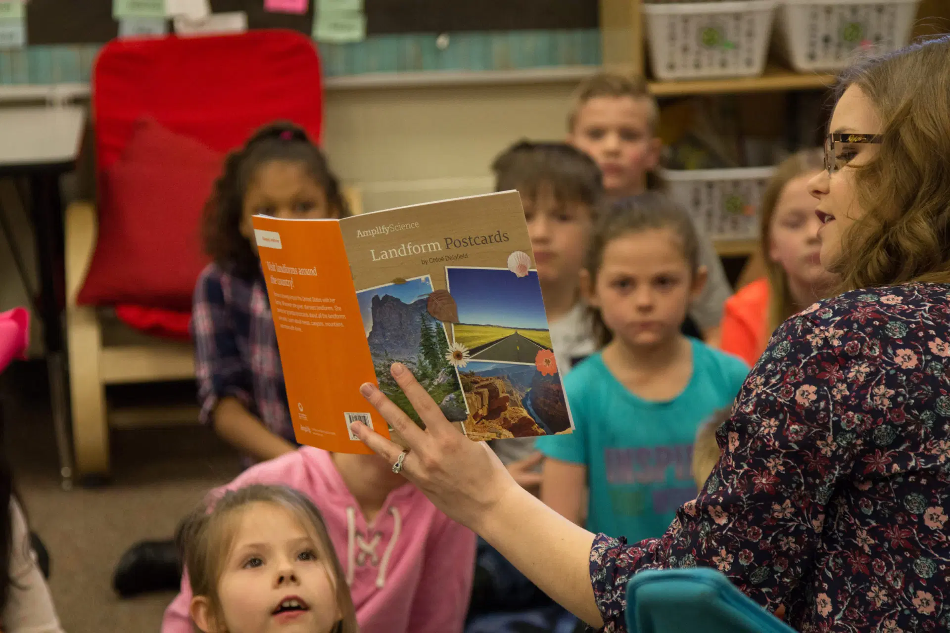 Una mujer lee un libro titulado "Postales de accidentes geográficos" a un grupo de niños atentos sentados en el suelo de un salón de clases.