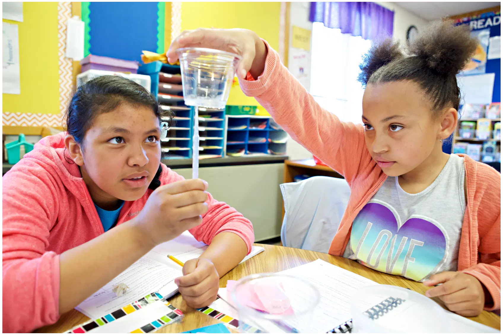 Dos niñas en un salón de clases realizan un experimento científico con agua y una pajita, una sostiene una taza y la otra observa. Las hojas de trabajo y los materiales están sobre la mesa.