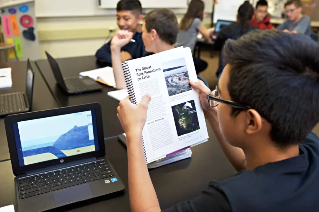 Estudiantes sentados en una mesa negra en un salón de clases, un estudiante lee un libro de texto sobre formaciones rocosas en la Tierra mientras otros discuten y trabajan en computadoras portátiles.