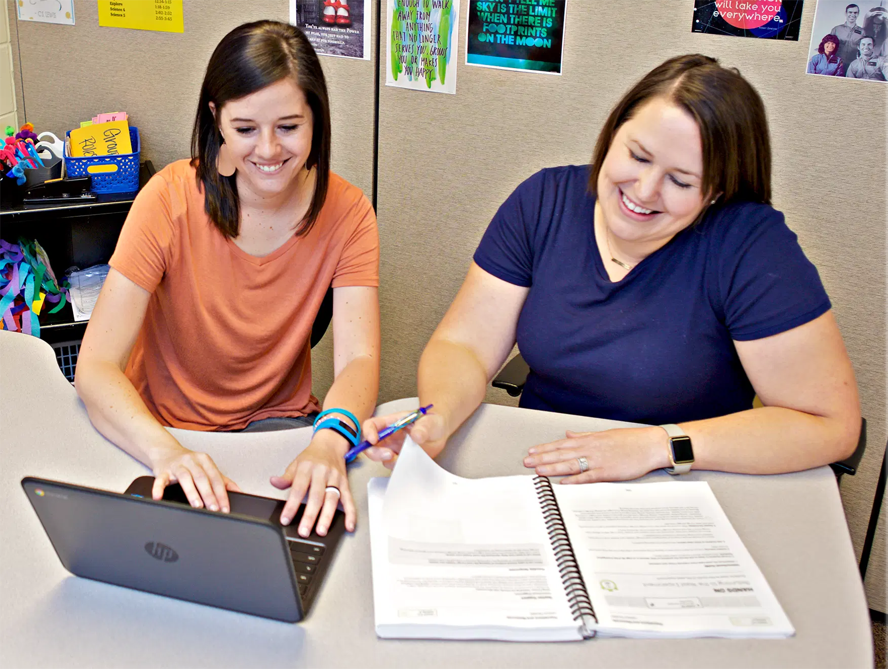 Two women sitting at a table. One is using a laptop, and the other is writing in a notebook. Both are smiling.