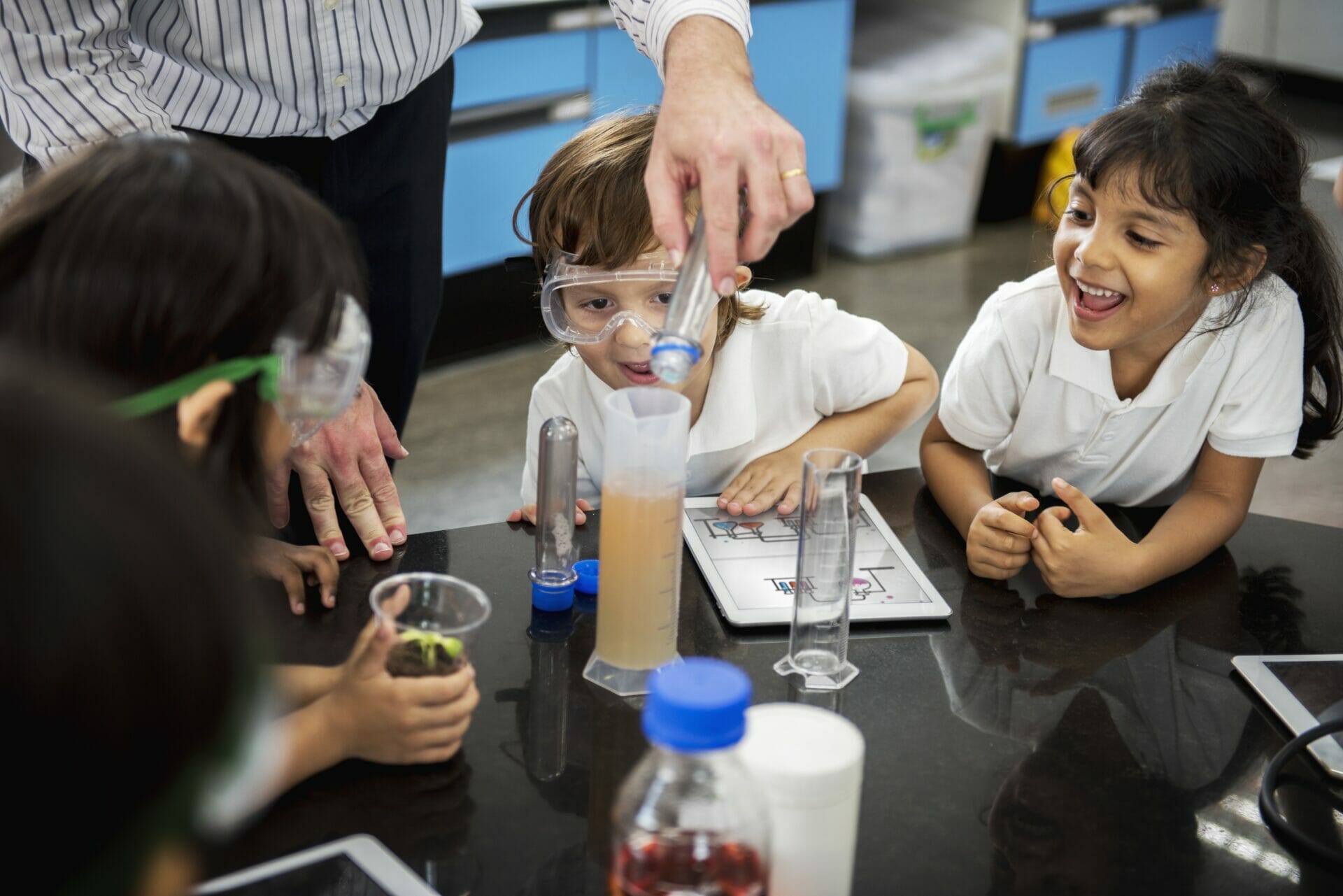 Los niños que usan gafas de seguridad participan en un experimento científico con un maestro, observando cómo se vierte un líquido en un cilindro graduado.