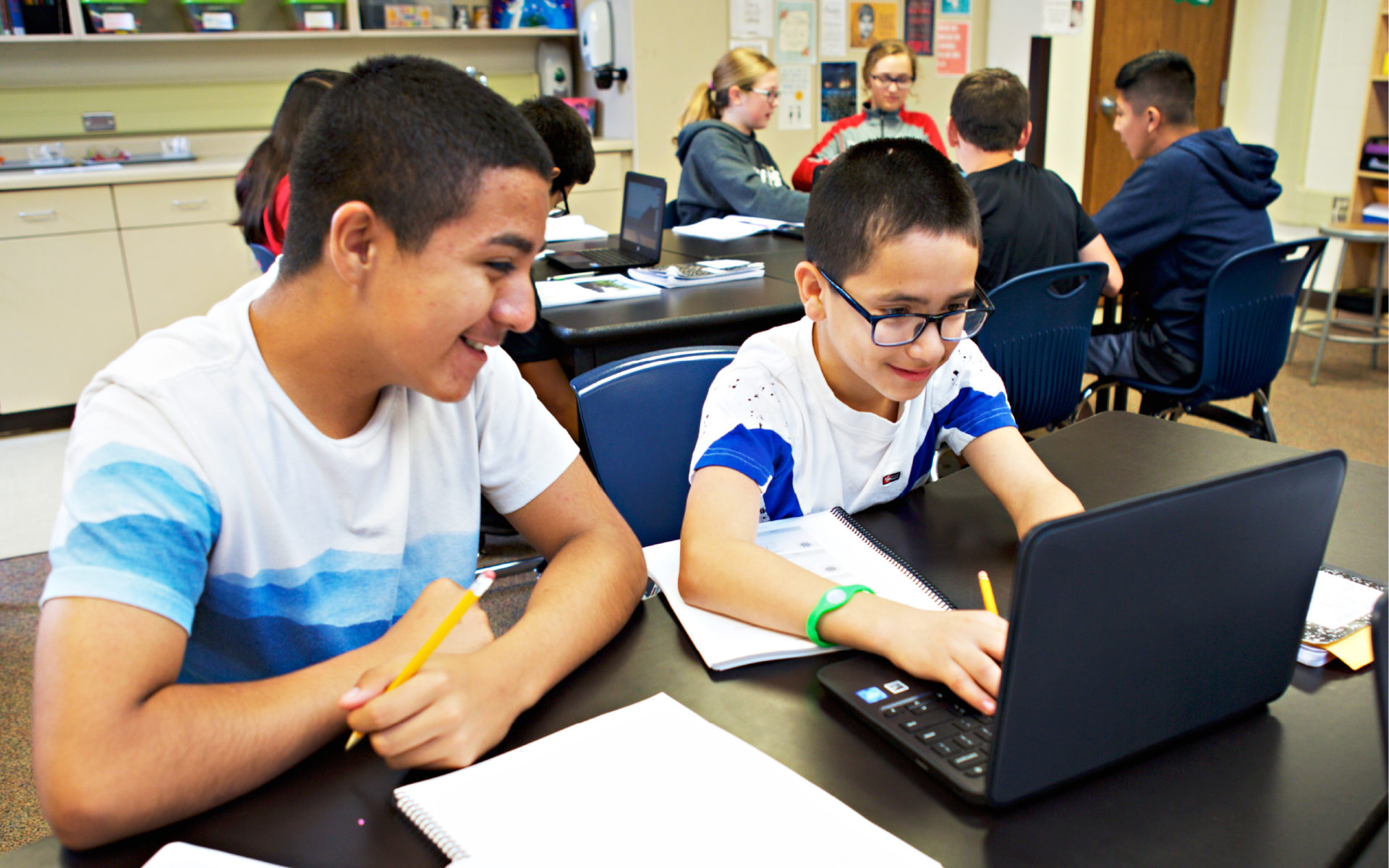 Two boys working on a laptop in a classroom, surrounded by other students and desks. They are smiling and writing on notebooks, appearing focused on the task.