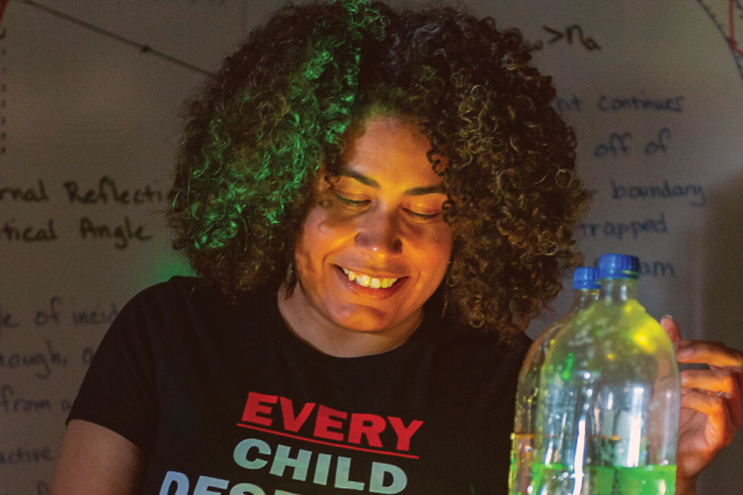 Person smiling while examining a plastic bottle with colorful lighting, standing in front of a whiteboard with text.