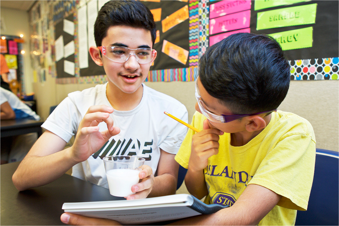 Two boys wearing safety glasses sit at a table. One holds a cup of liquid and gestures, while the other holds a pencil and notebook, engaged in discussion.