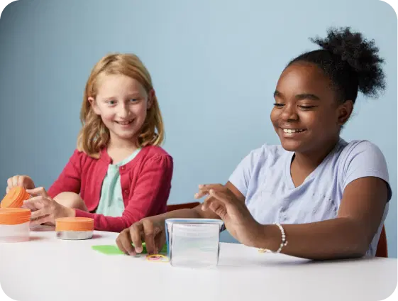 Two girls sit at a table, smiling as they explore colorful craft materials, including small containers and papers. Against a light blue background, their project takes shape, reflecting the curious spirit of middle school science enhanced by Amplify Science.