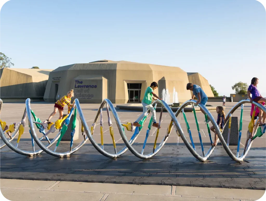 Children enthusiastically climbing a DNA sculpture at The Lawrence Hall of Science, echoing the spirit of Amplify Science, with the building and a bubbling fountain providing a captivating backdrop that inspires young minds to delve deeper into middle school science wonders.