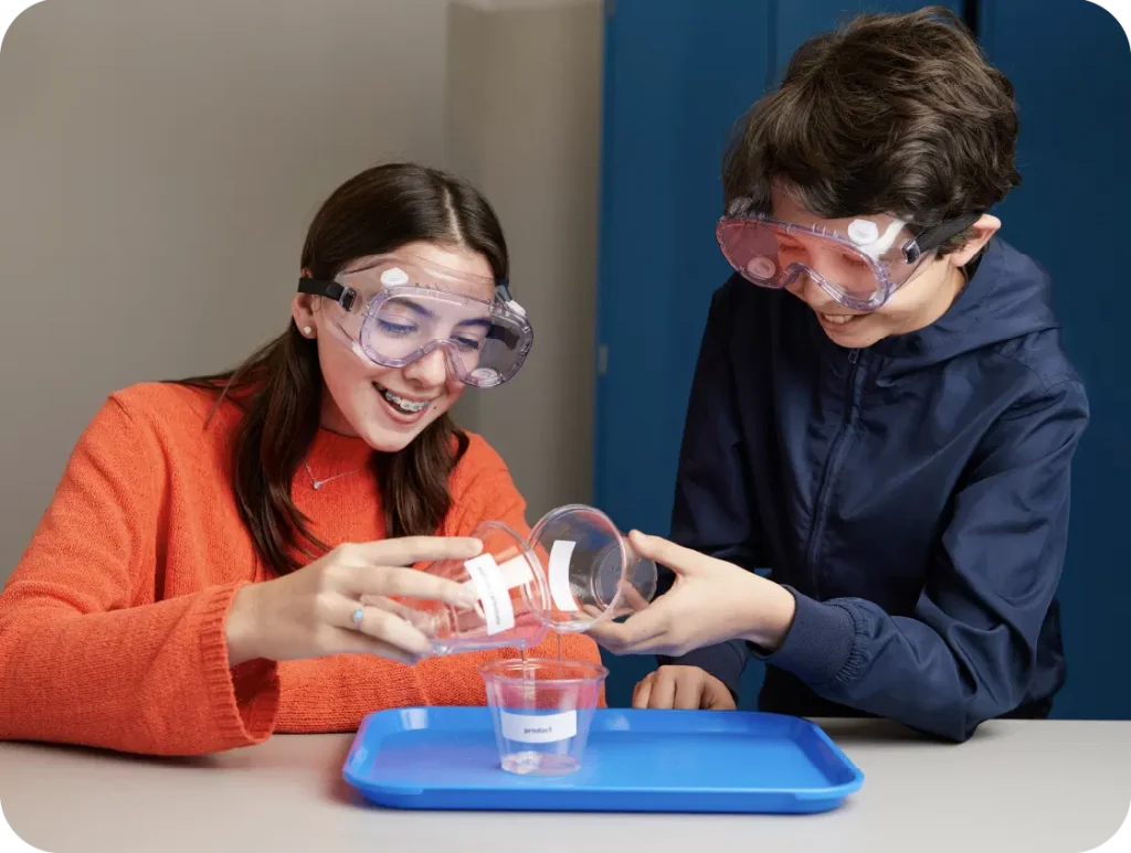 Two kids in safety goggles pour liquid from a bottle into a clear container over a blue tray, fully engaged in an Amplify Science activity. Their curiosity and excitement shine as they explore the wonders of middle school science.