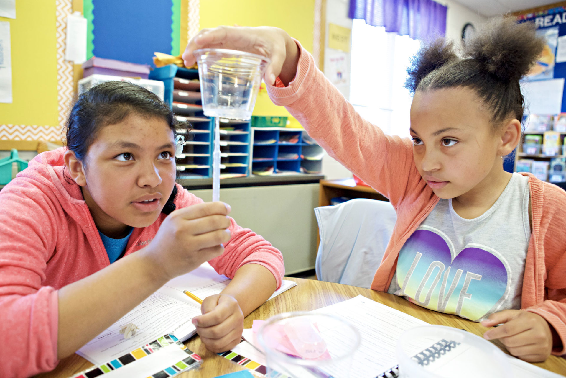 Two students in a classroom examine a cup of water held by one student. Both are focused on the experiment at their desk.
