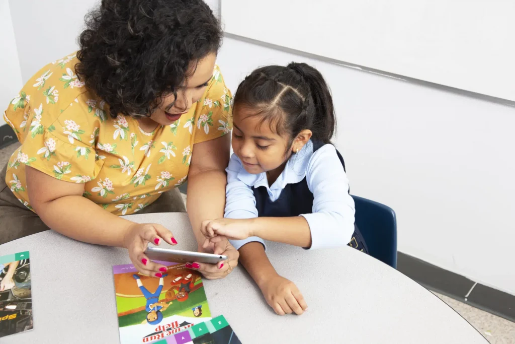 A woman and a young girl are sitting at a table looking at a smartphone together. A children's book and a magazine are on the table.