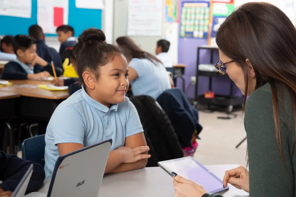 Teacher and student sitting across from each other in a classroom, with the teacher holding a tablet. Other students are working in the background.