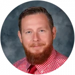 A person with a reddish-brown beard and short hair wearing a red and white checkered shirt and red tie against a gray background.