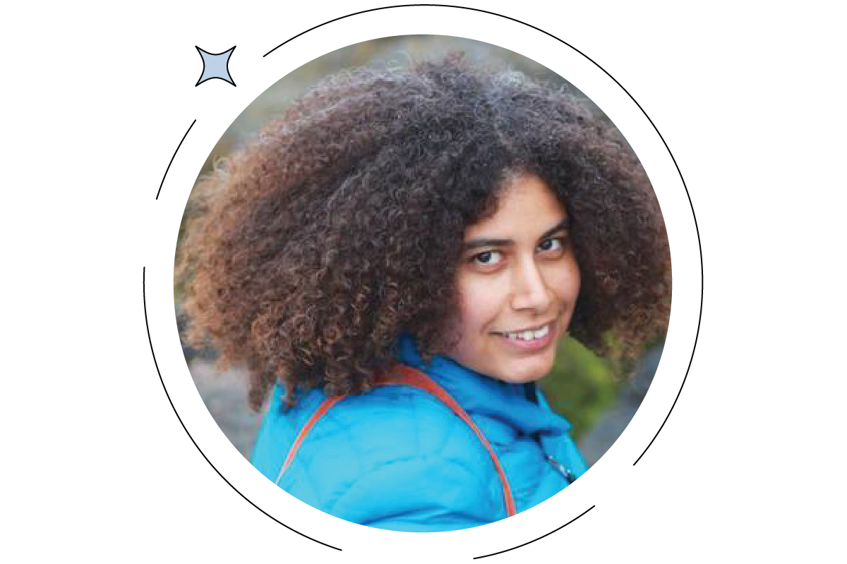Person with curly hair wearing a blue jacket, smiling at the camera with a blurred background.
