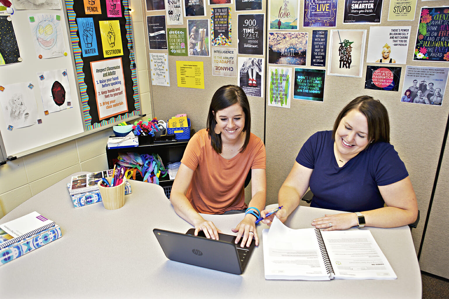 Two women sitting at a table working on a laptop and documents, surrounded by colorful posters on the walls.