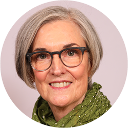 An older adult with short gray hair and glasses, wearing a green scarf, smiles directly at the camera.