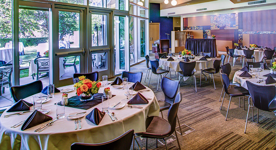Banquet room set for an event with round tables, black chairs, white tablecloths, black napkins, and floral centerpieces. Large windows offer a view of a garden outside.