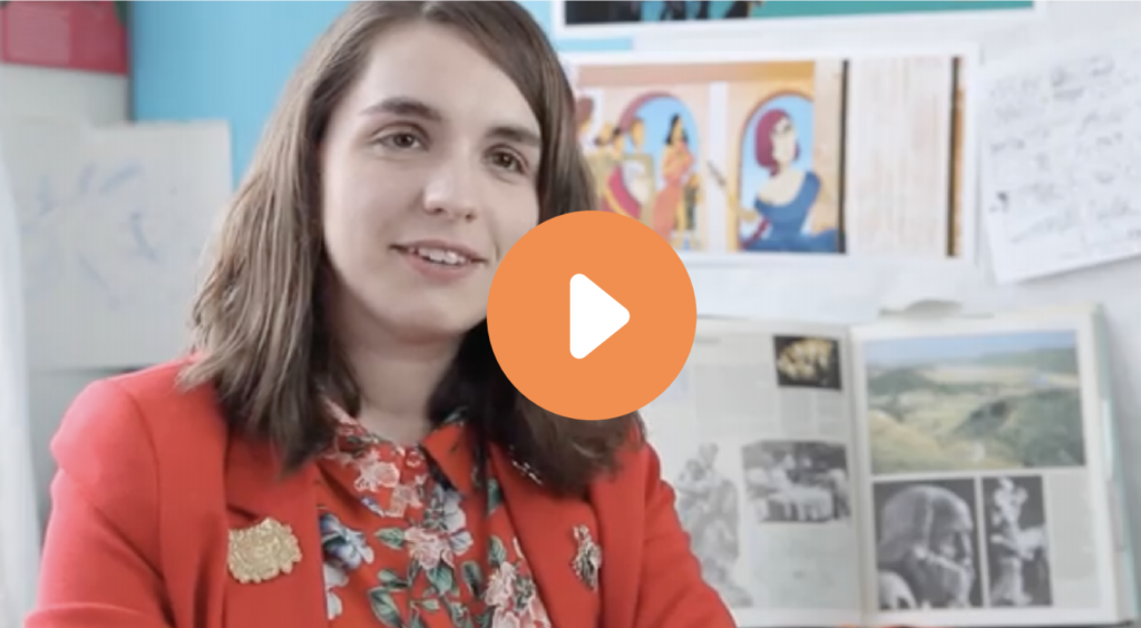 Person wearing a red jacket and floral blouse sits in front of a wall with artwork and notes, partially obscured by a large play button icon.