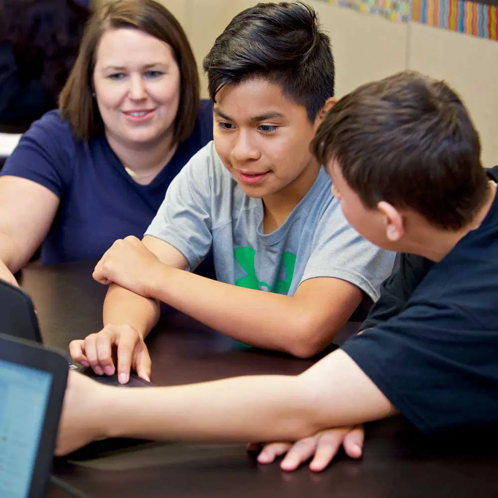 Two boys work together on a laptop while a woman—likely one of their middle school teachers—sits beside them, observing and smiling in a classroom setting.
