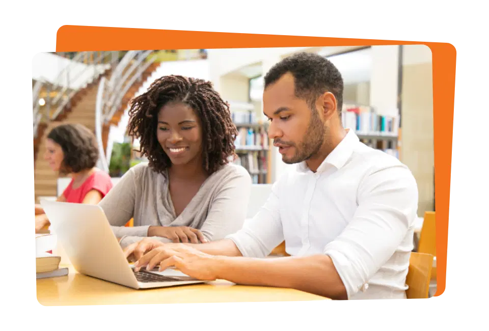 Two people at a library table work together on a laptop, with shelves and a spiral staircase in the background.