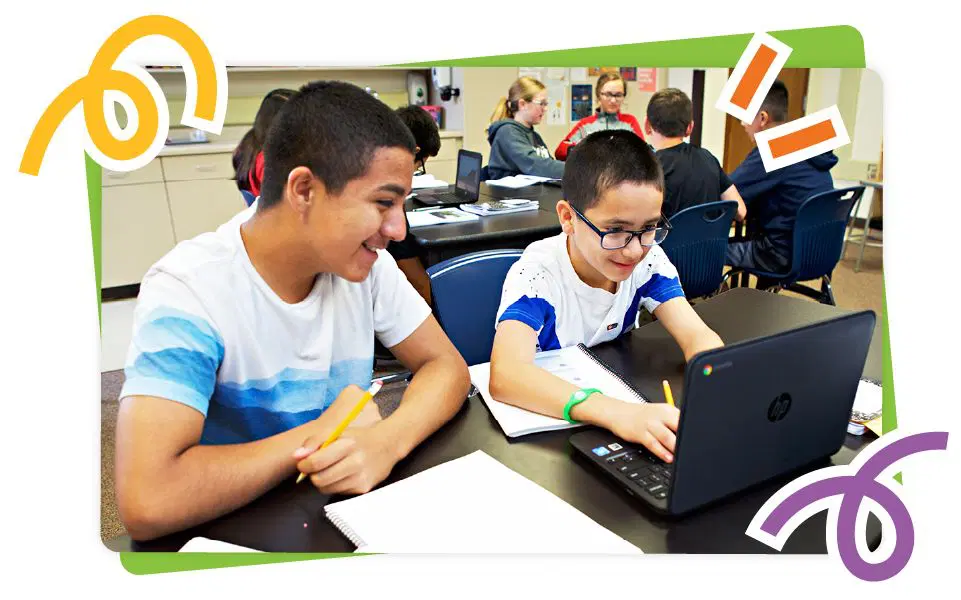 Two boys in a classroom working on a laptop together, surrounded by notebooks and other students in the background.
