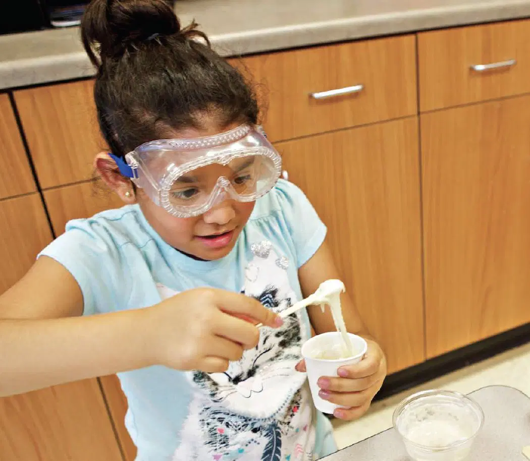A young girl wearing safety goggles stretches a gooey substance from a cup in a kitchen or classroom setting.