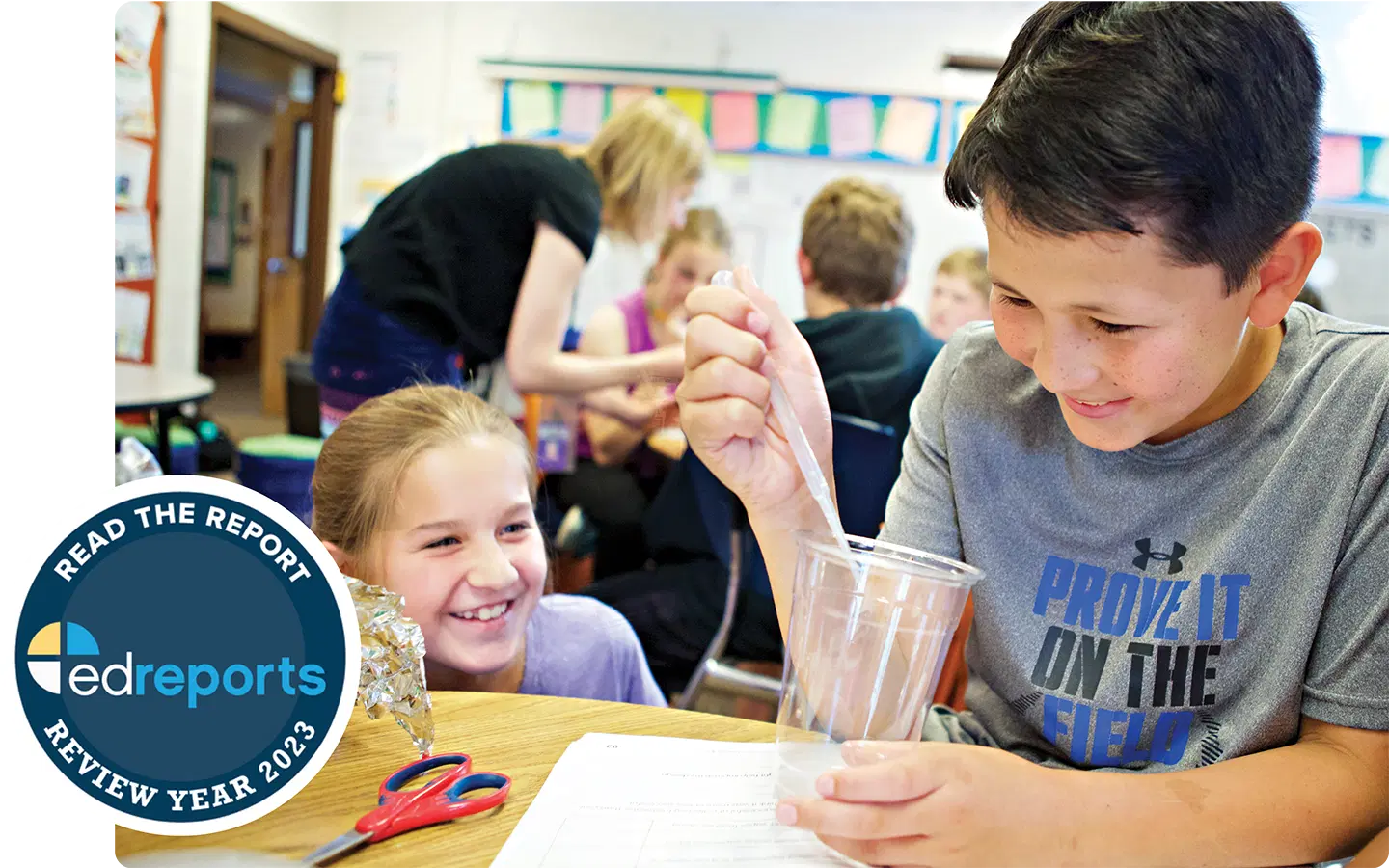 A boy stirs a clear liquid in a plastic cup while a girl smiles beside him in a classroom with students and a teacher in the background. EdReports badge is overlaid in the corner.