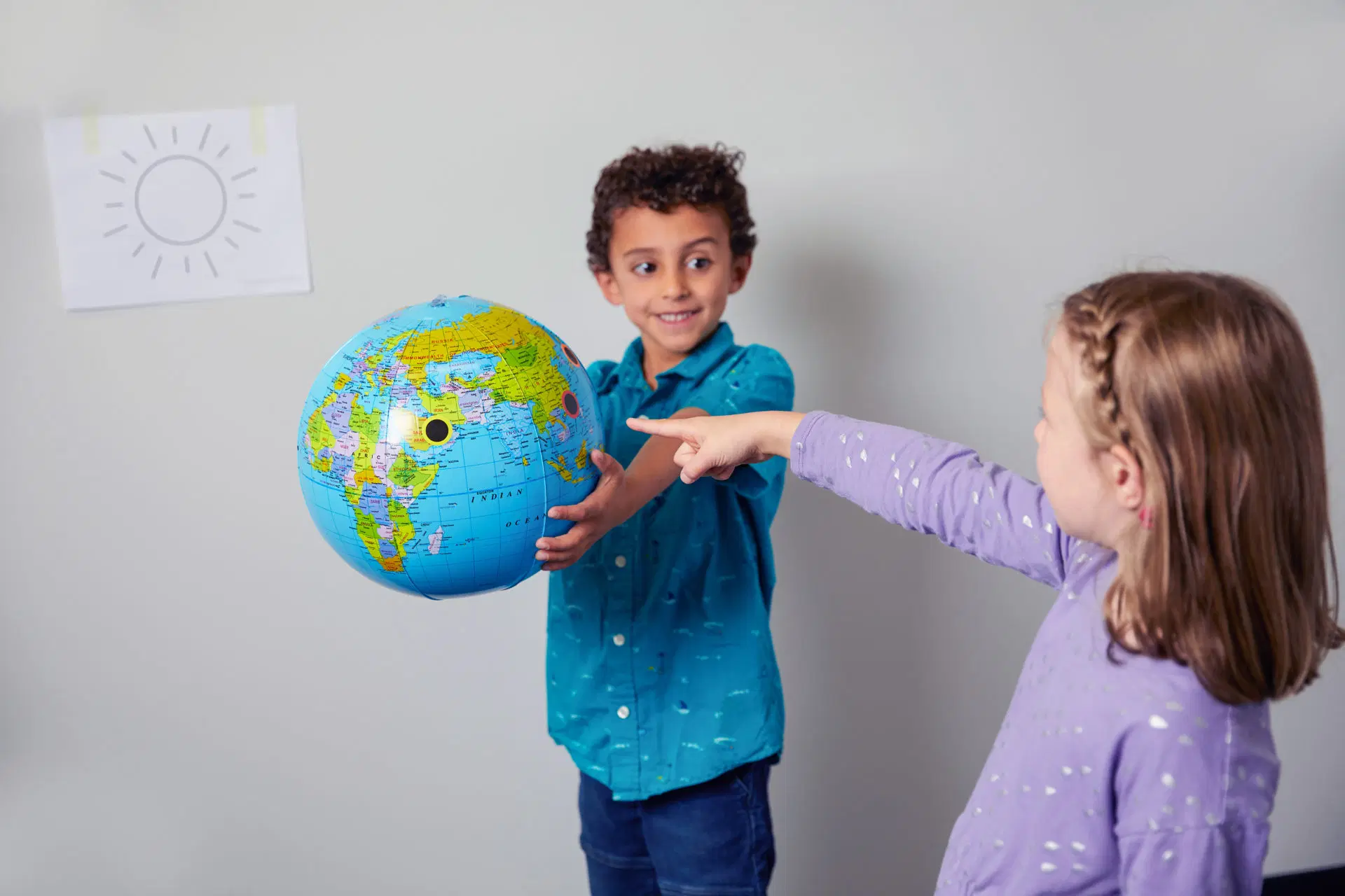 Two children stand indoors; one holds a globe while the other points at it. A drawing of a sun is taped to the wall in the background.