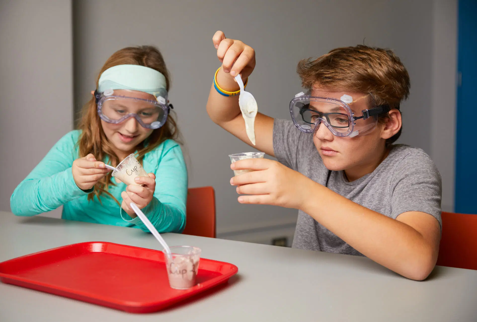 Two children wearing safety goggles conduct a science experiment, using plastic spoons to mix substances in clear cups at a table with a red tray.