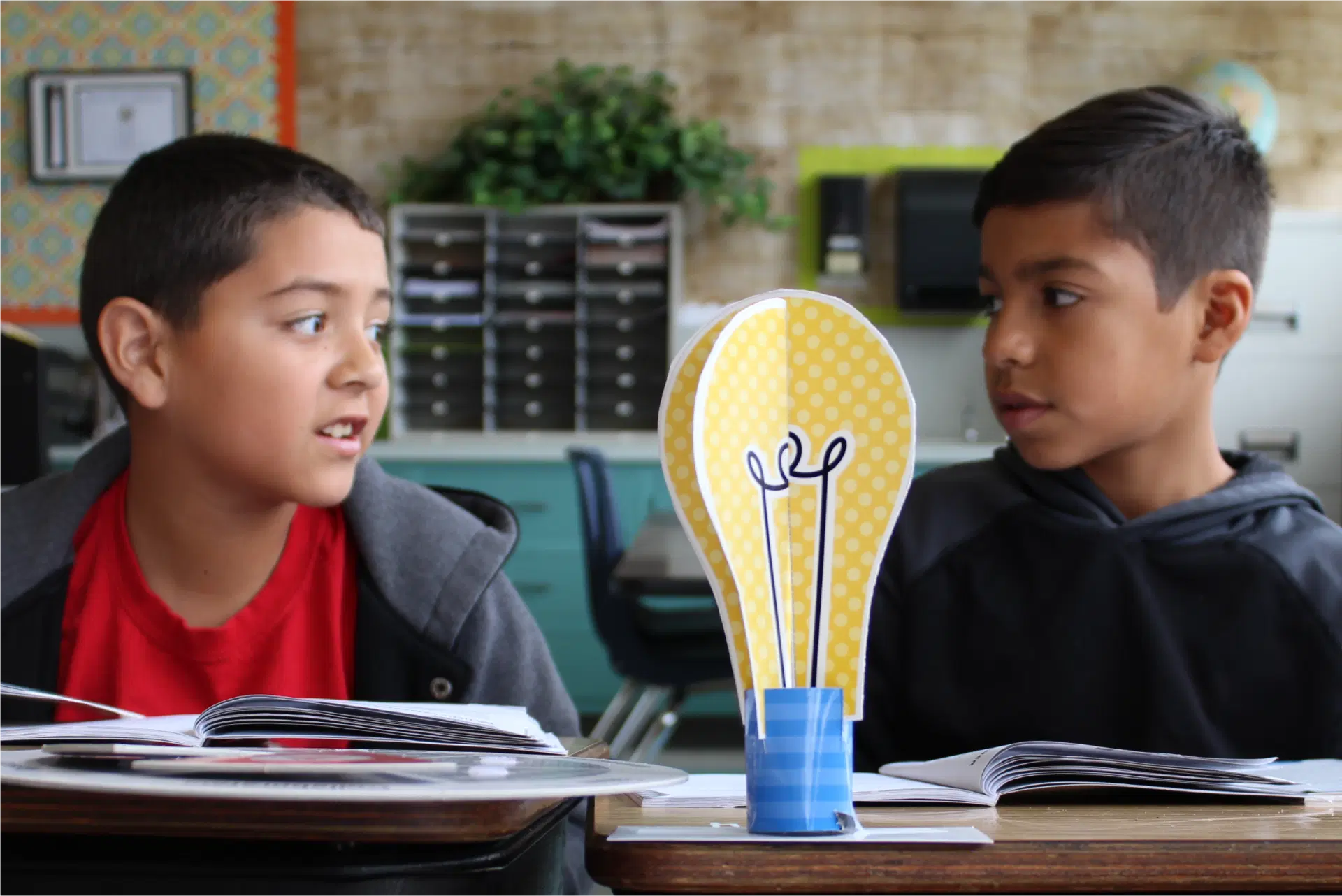 Two boys sit at desks with open books, talking to each other. A paper model shaped like a lightbulb stands between them in a classroom setting.