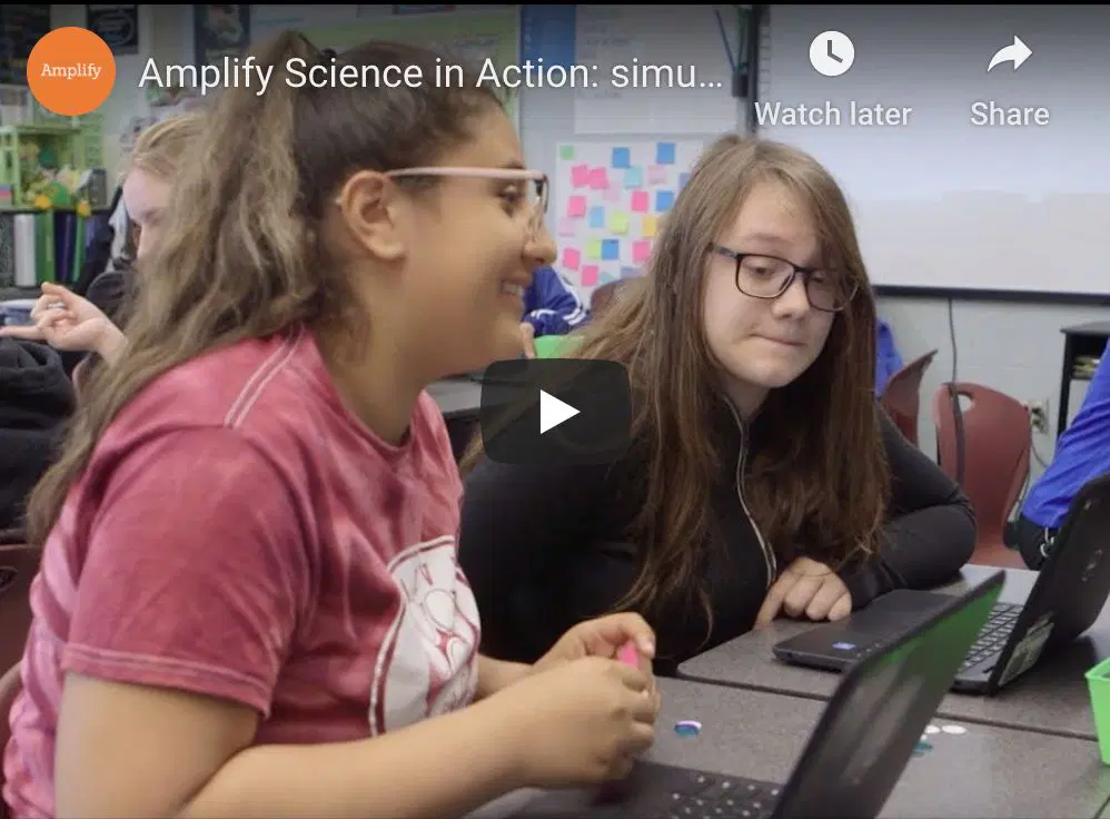 Two students sit at a table in a classroom, working on laptops and talking, with other students visible in the background.