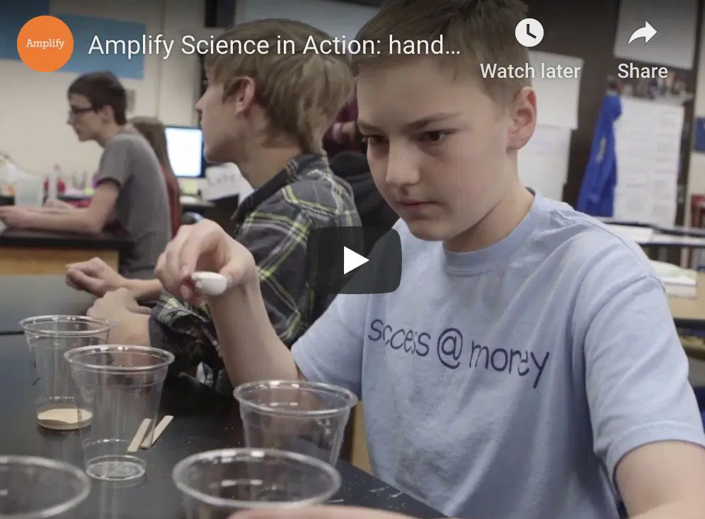 A student in a classroom uses a dropper to add liquid to clear plastic cups during a science experiment. Other students and lab materials are visible in the background.