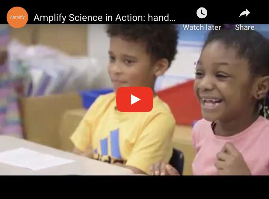 Two young children sit at a classroom table, smiling and engaged, with a large red play button overlayed, indicating a paused YouTube video.