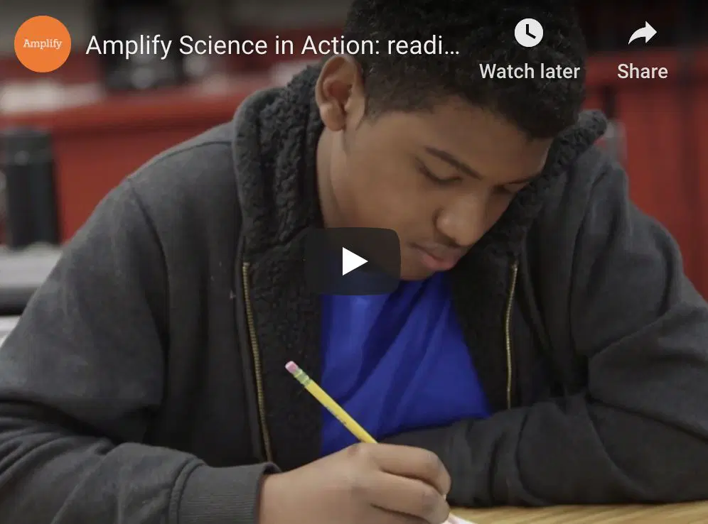 A student sits at a desk writing with a pencil while focused on his work, with classroom materials visible in the background.