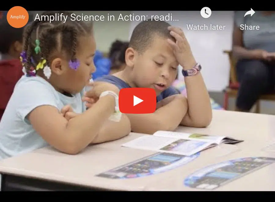 Two young children sit at a classroom table reading a book together, with papers spread out in front of them.