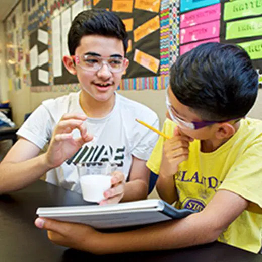 Two boys wearing safety glasses sit at a table; one holds a cup with liquid while the other holds a pencil and notebook. Classroom materials are visible in the background.